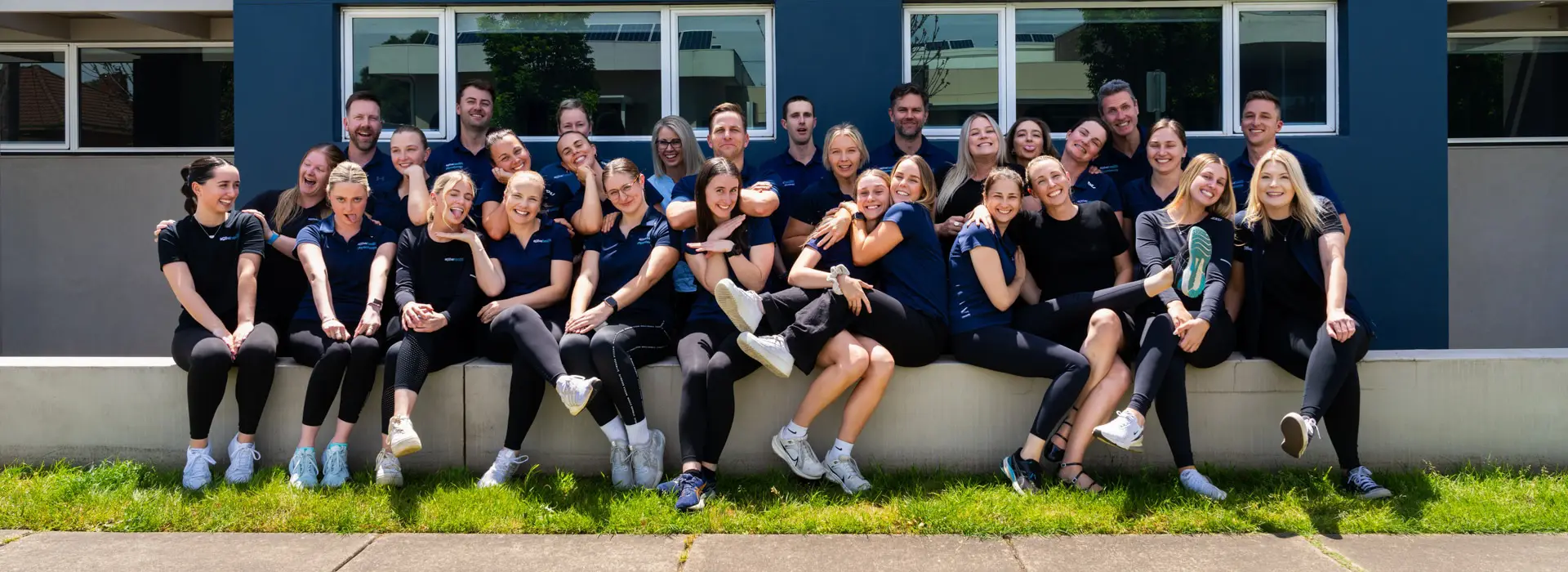 Group of 27 people smiling and posing in front of a building with blue and beige walls, sitting and standing on a low concrete ledge.