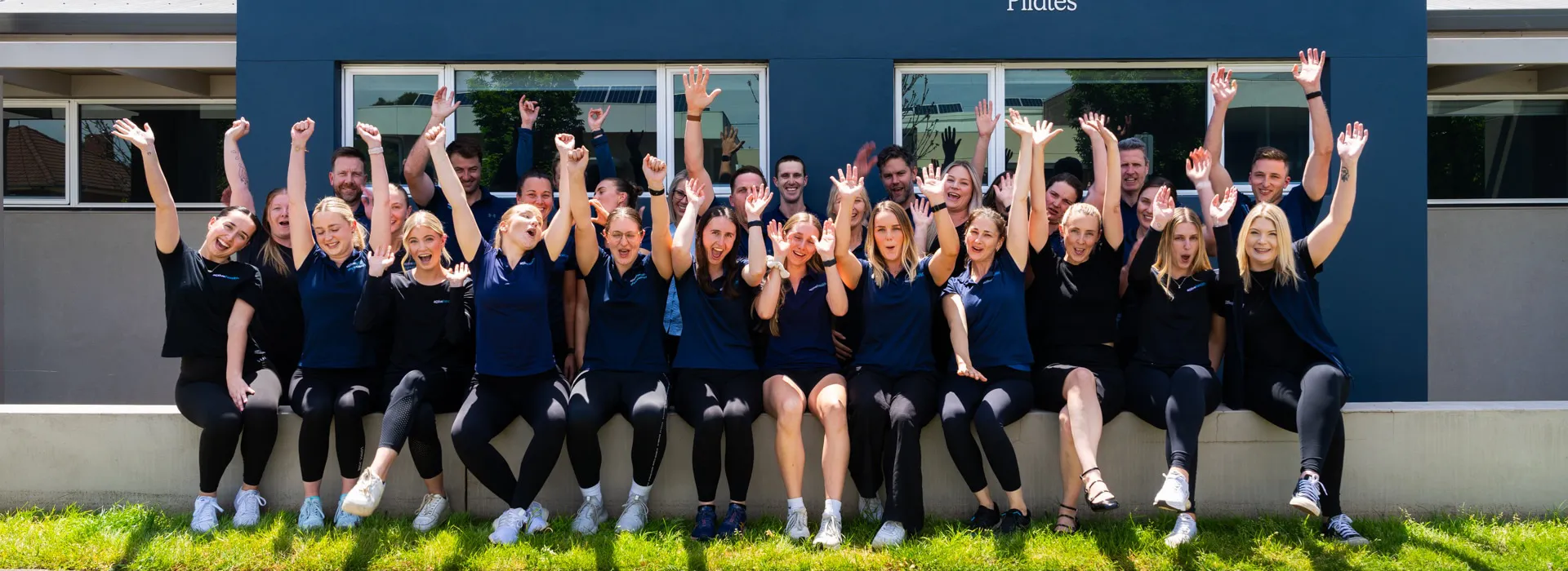 Group of 25 people sitting outdoors on a low wall, smiling and raising their hands enthusiastically.
