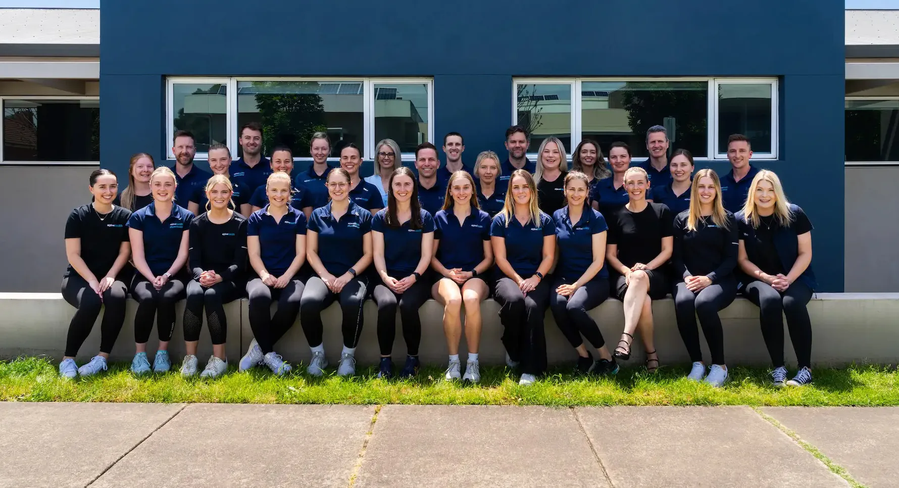 Group portrait of 28 people in matching active health uniforms seated and standing in front of a blue building wall with windows.
