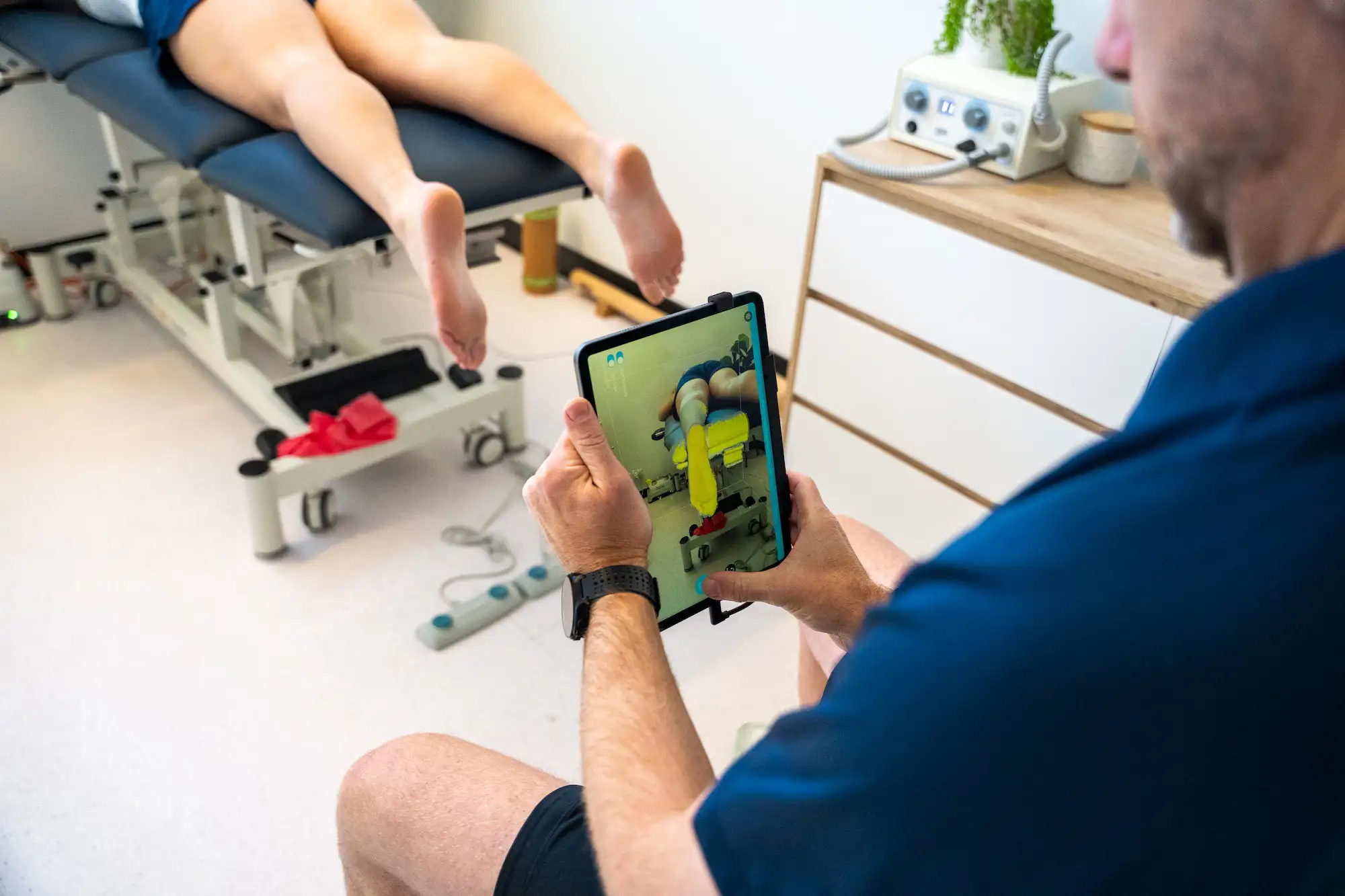 Man holding a tablet showing a therapy session with a person lying face down on a treatment table in a clinical setting.