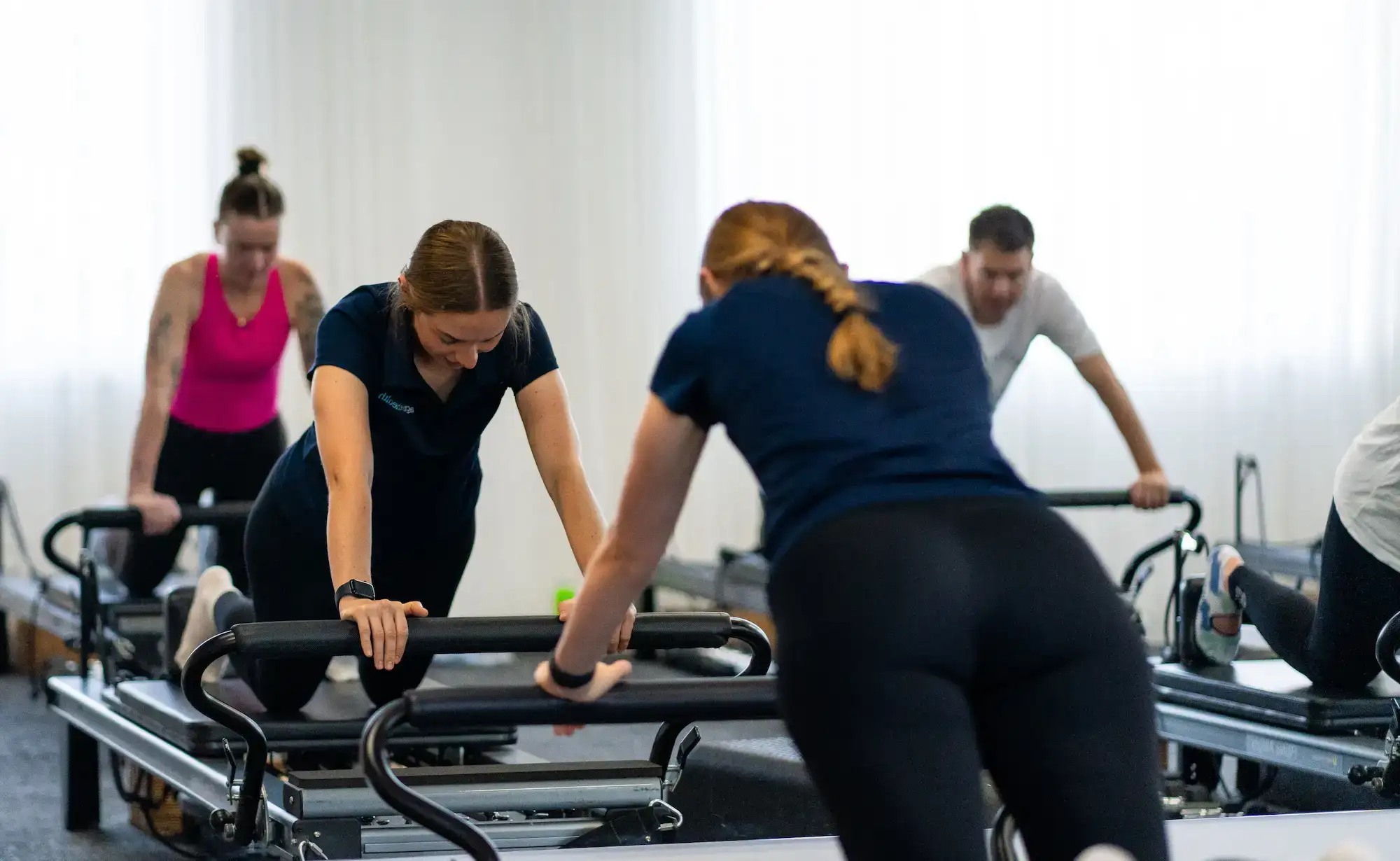 Group of adults exercising on Pilates reformer machines in a bright studio.