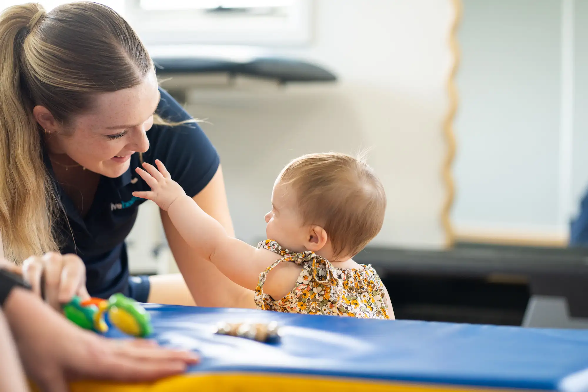 A smiling woman interacts with a baby who reaches out with their hand while sitting on a blue mat.