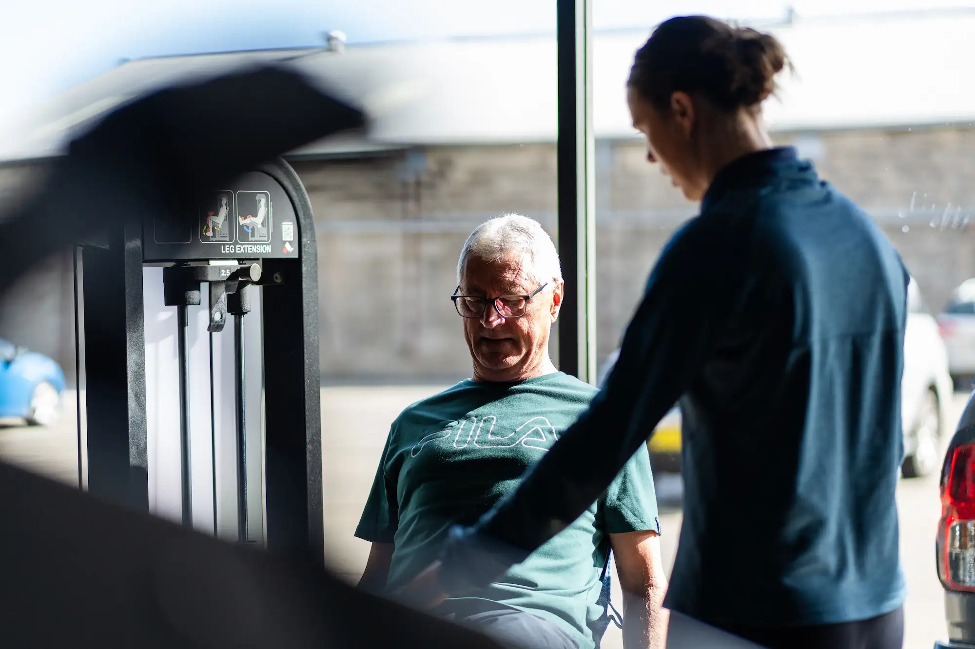 Older man wearing glasses and a green FILA shirt using a leg extension machine at a gym, assisted by a trainer.