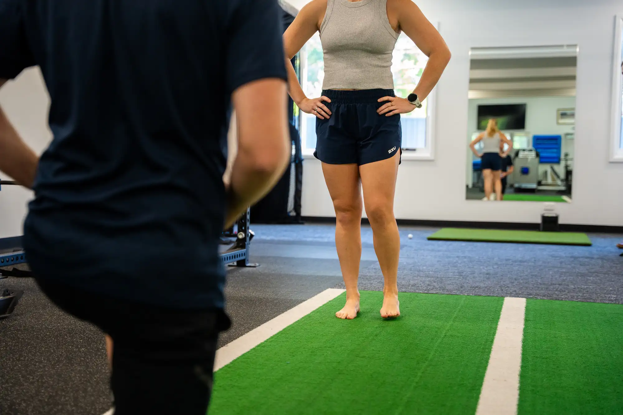 Woman standing barefoot on a green exercise mat indoors with hands on hips, facing a trainer in a gym setting.