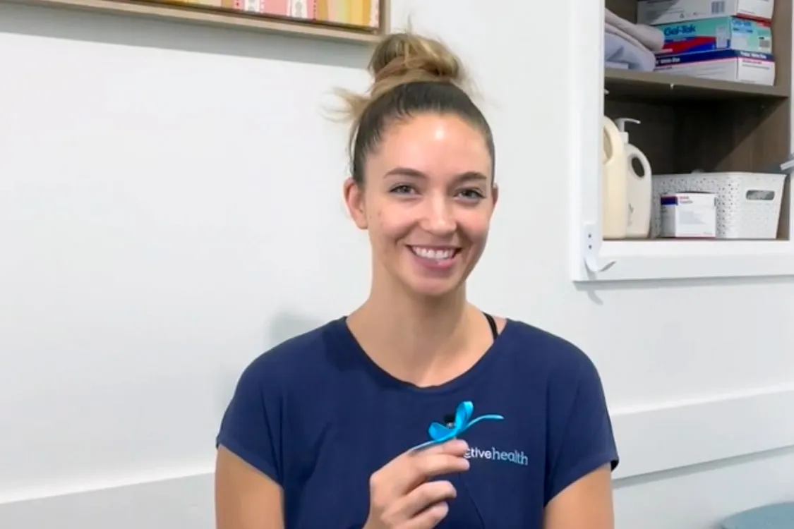 Smiling woman with hair in a bun wearing a navy shirt holding a blue ribbon, sitting indoors near a cabinet with medical supplies.