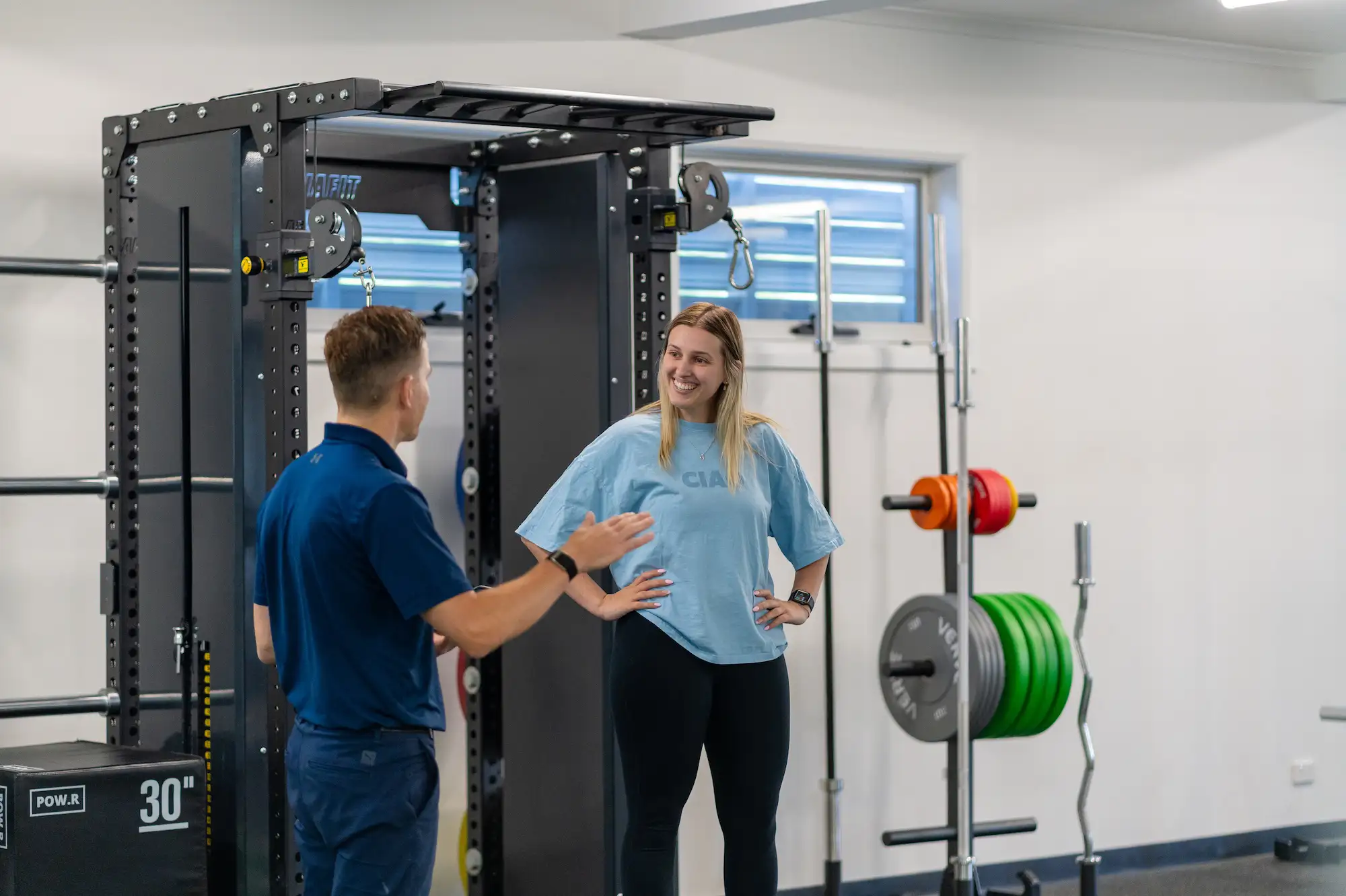 A woman in workout clothes smiling and talking with a man in a blue shirt inside a gym with exercise equipment.