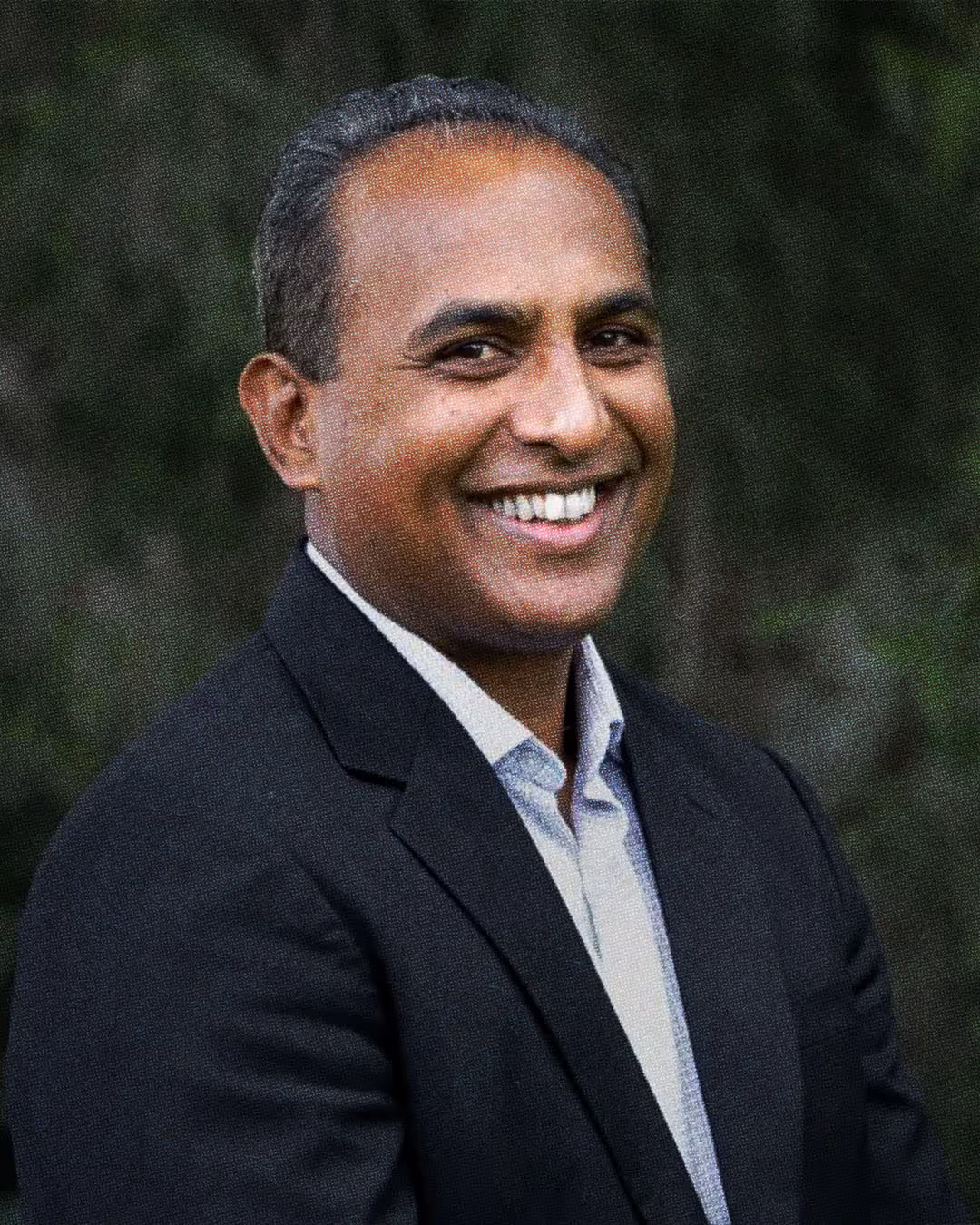 Smiling man with short hair wearing a dark blazer and light-colored shirt standing against a green blurred background.