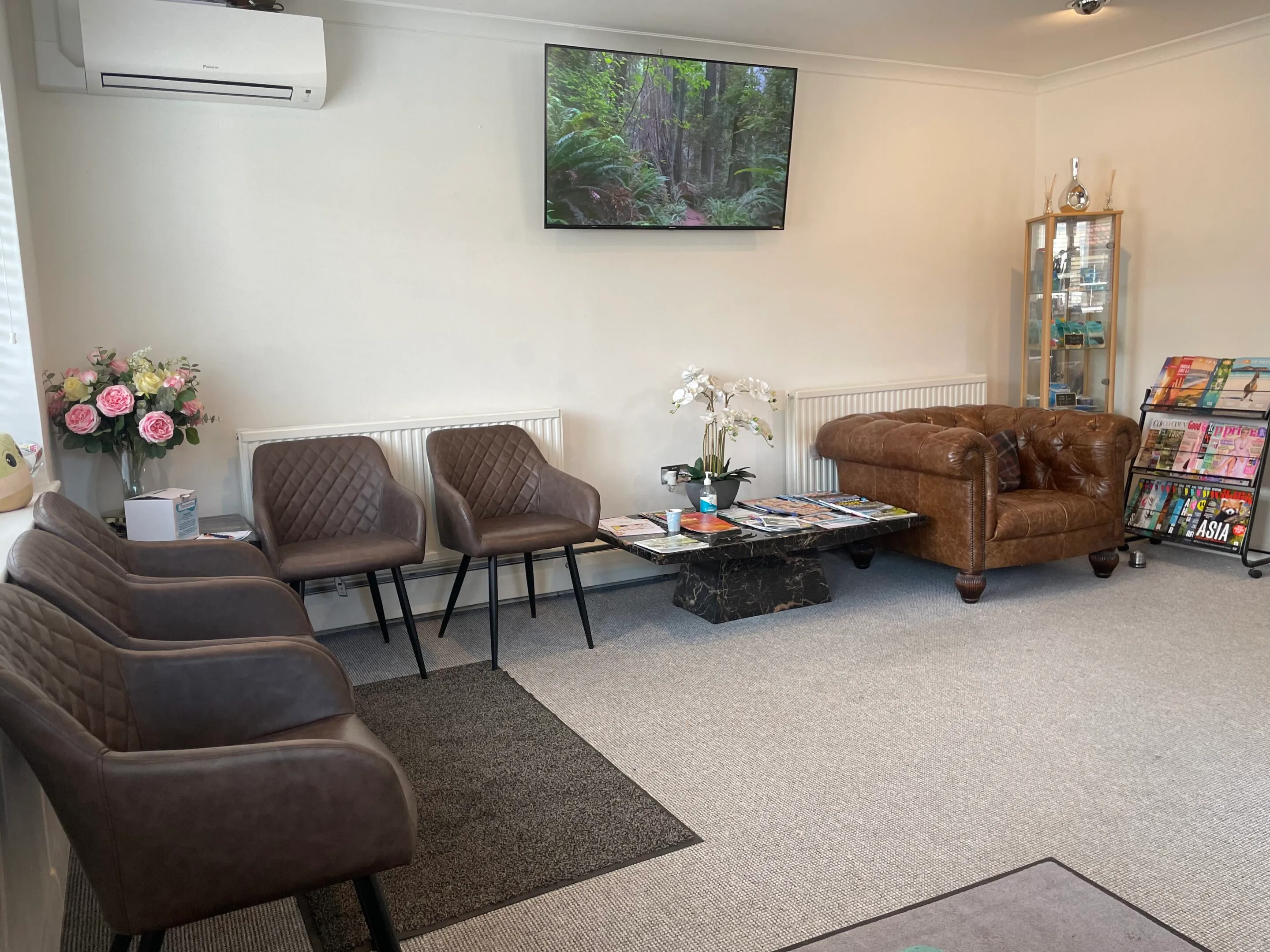 Modern waiting room with brown leather chairs and sofa, a marble coffee table with magazines, flowers, and a wall-mounted TV displaying a forest scene.