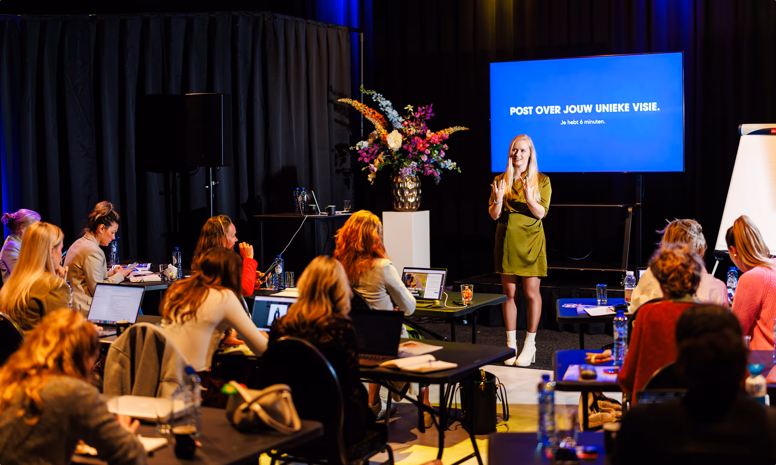 Een vrouw in een groene jurk geeft een presentatie aan een groep mensen in een zaal, met een scherm waarop staat 'POST OVER JOUW UNIEKE VISIE. Je hebt 6 minuten.'
