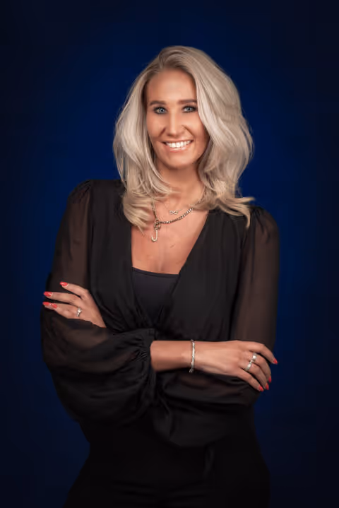 Blonde vrouw met golvend haar, zwart blouse, gekruiste armen en een vriendelijke glimlach tegen een donkerblauwe achtergrond.