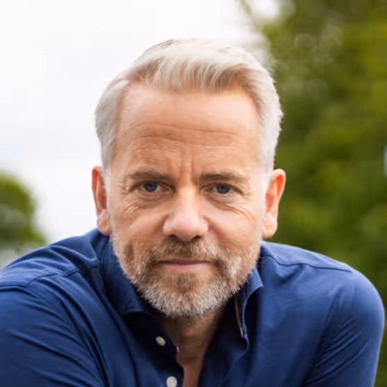 Close-up van een volwassen man met grijs haar en baard, gekleed in een blauwe blouse, zittend buiten met groen gebladerte op de achtergrond.