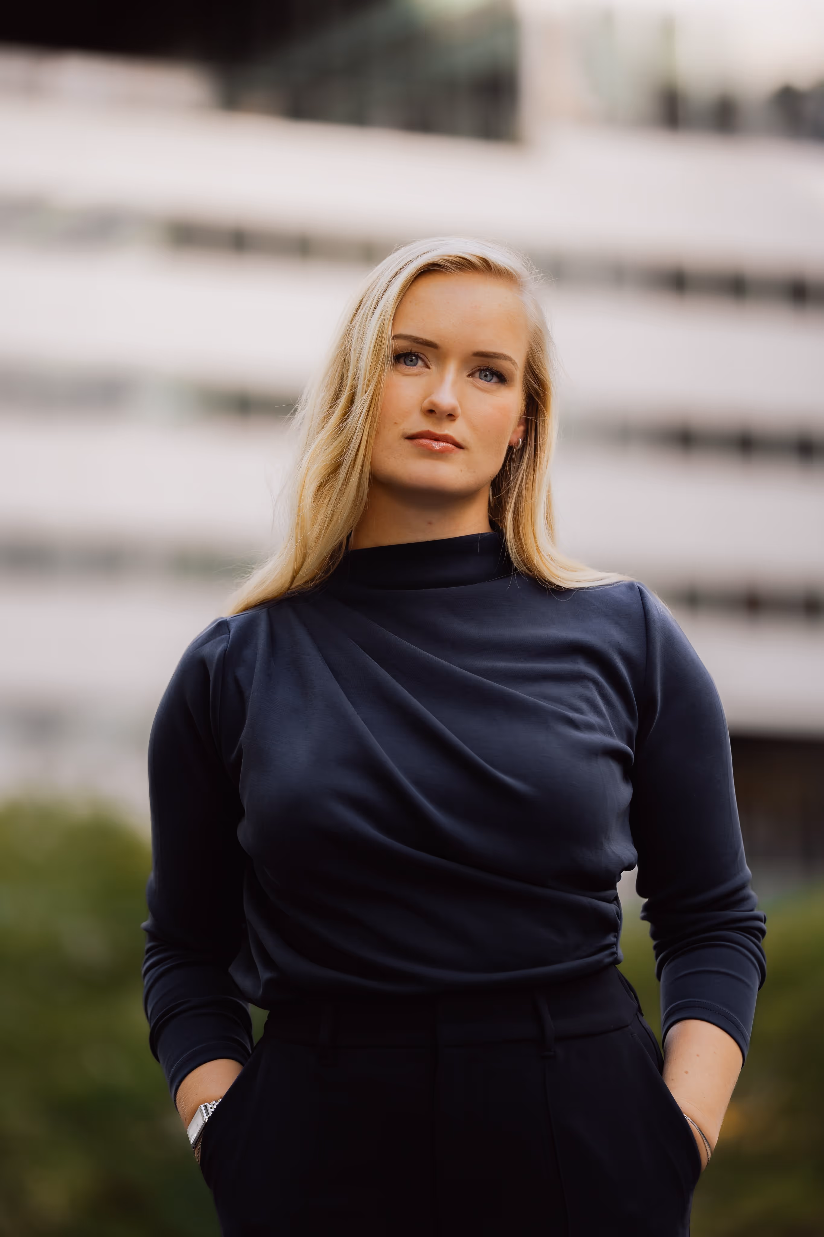 Blonde vrouw met blauwe ogen draagt een donkerblauwe trui en staat met haar handen in haar zakken buiten.