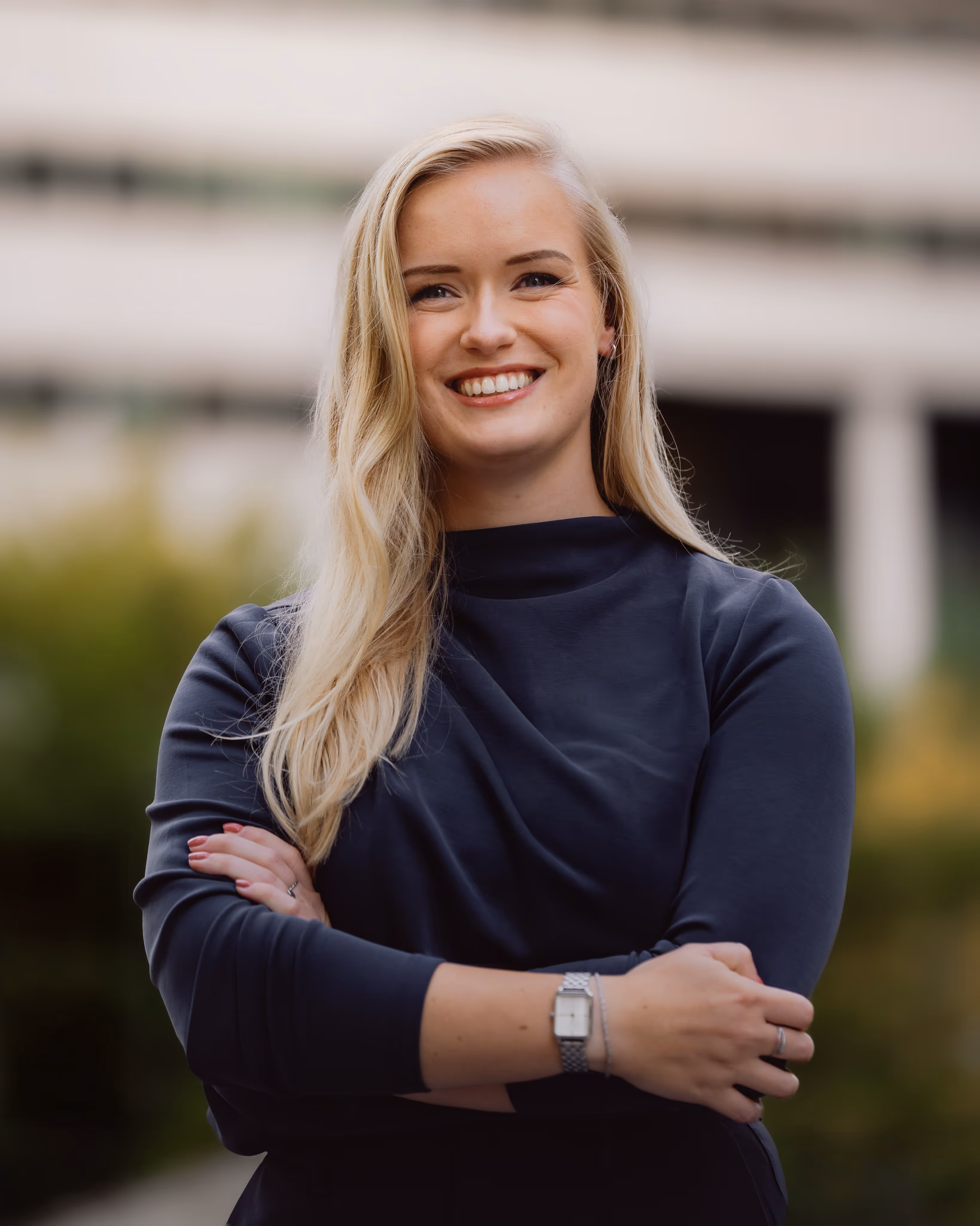 Blonde vrouw met lang haar en een zwarte trui staat buiten met armen gekruist en glimlacht.