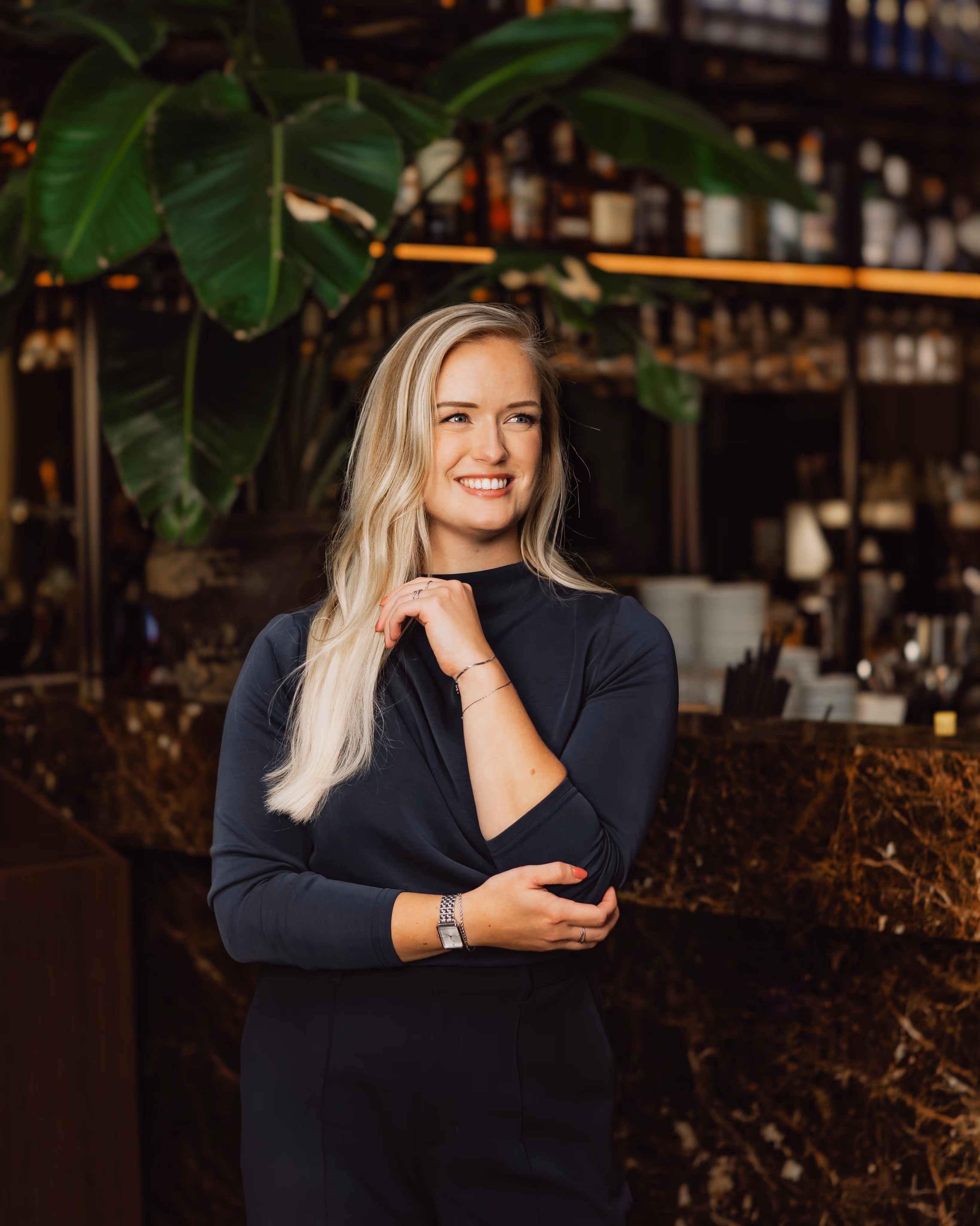 Blonde vrouw in een donkerblauwe trui glimlacht en staat met gekruiste armen voor een bar met planten op de achtergrond.