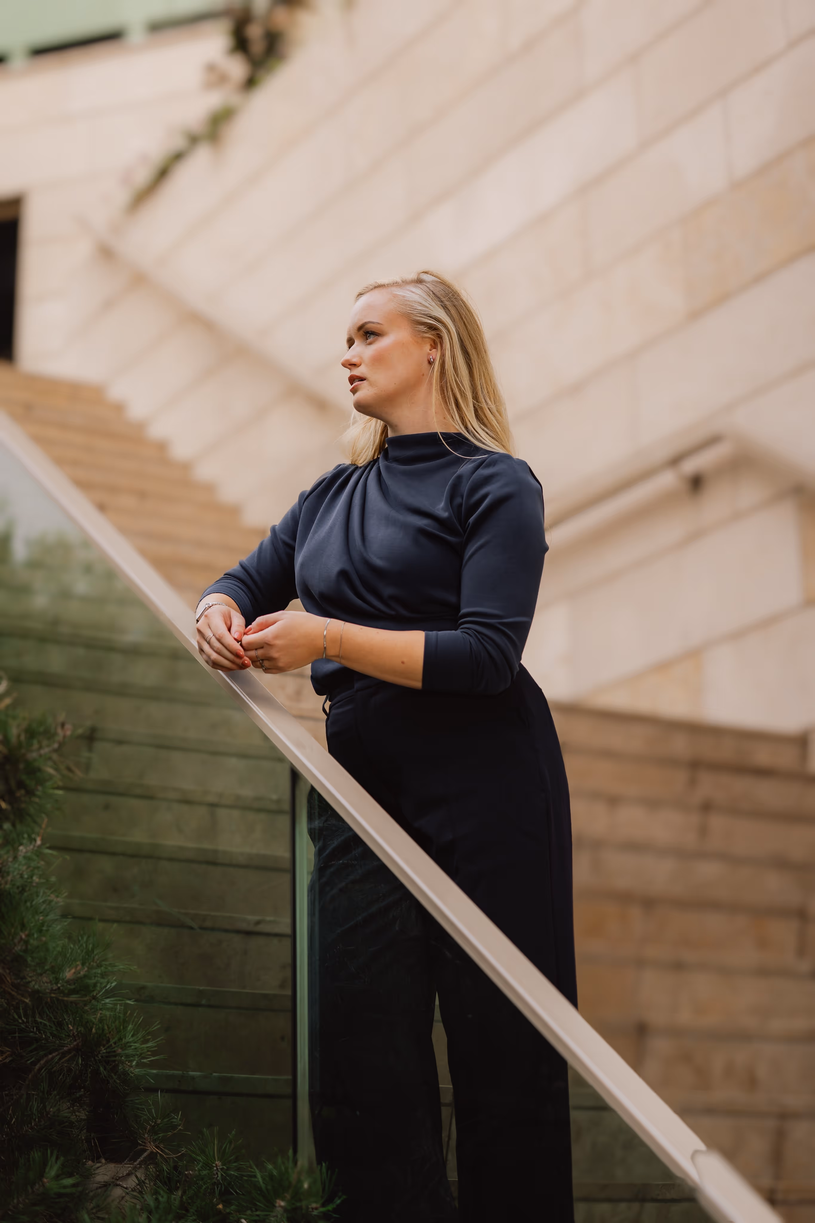 Vrouw met blonde haren leunt op een glazen balustrade langs een trap, kijkend naar links met een nadenkende uitdrukking.