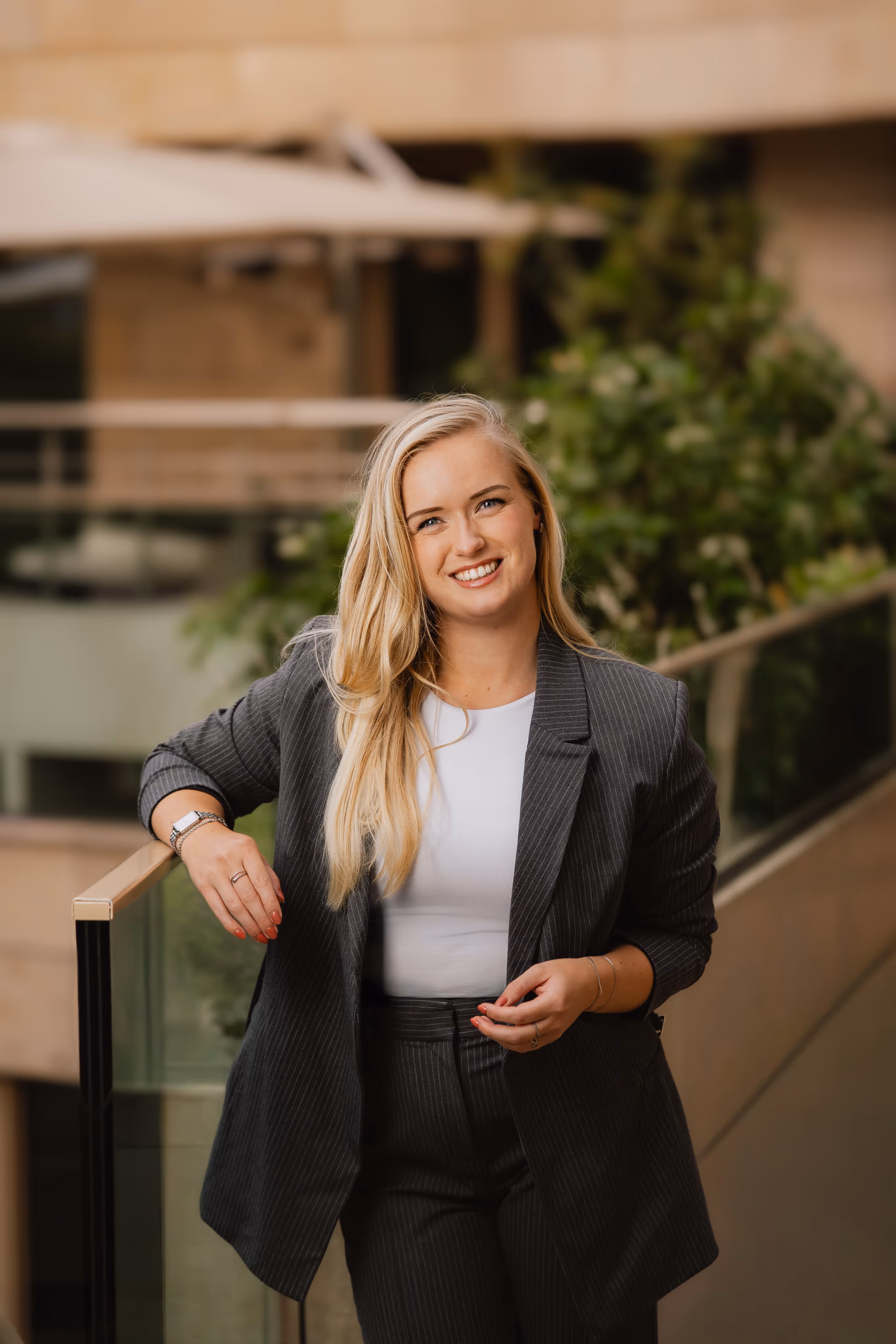 Vrouw met lang blond haar, gekleed in een zwart-pak met witte top, leunend tegen een glazen balustrade met groenblijvende planten op de achtergrond.