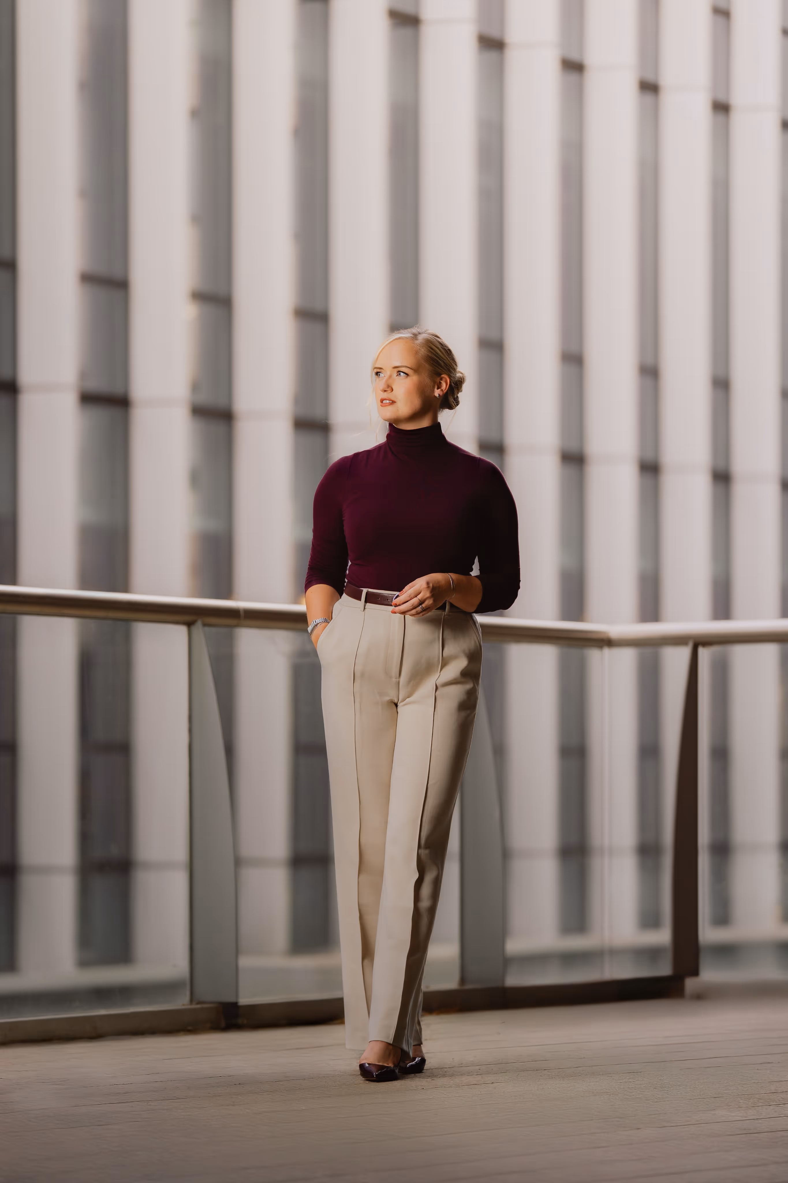Vrouw in een bordeauxrode coltrui en beige broek staat op een balkon met glazen balustrade voor een modern gebouw.