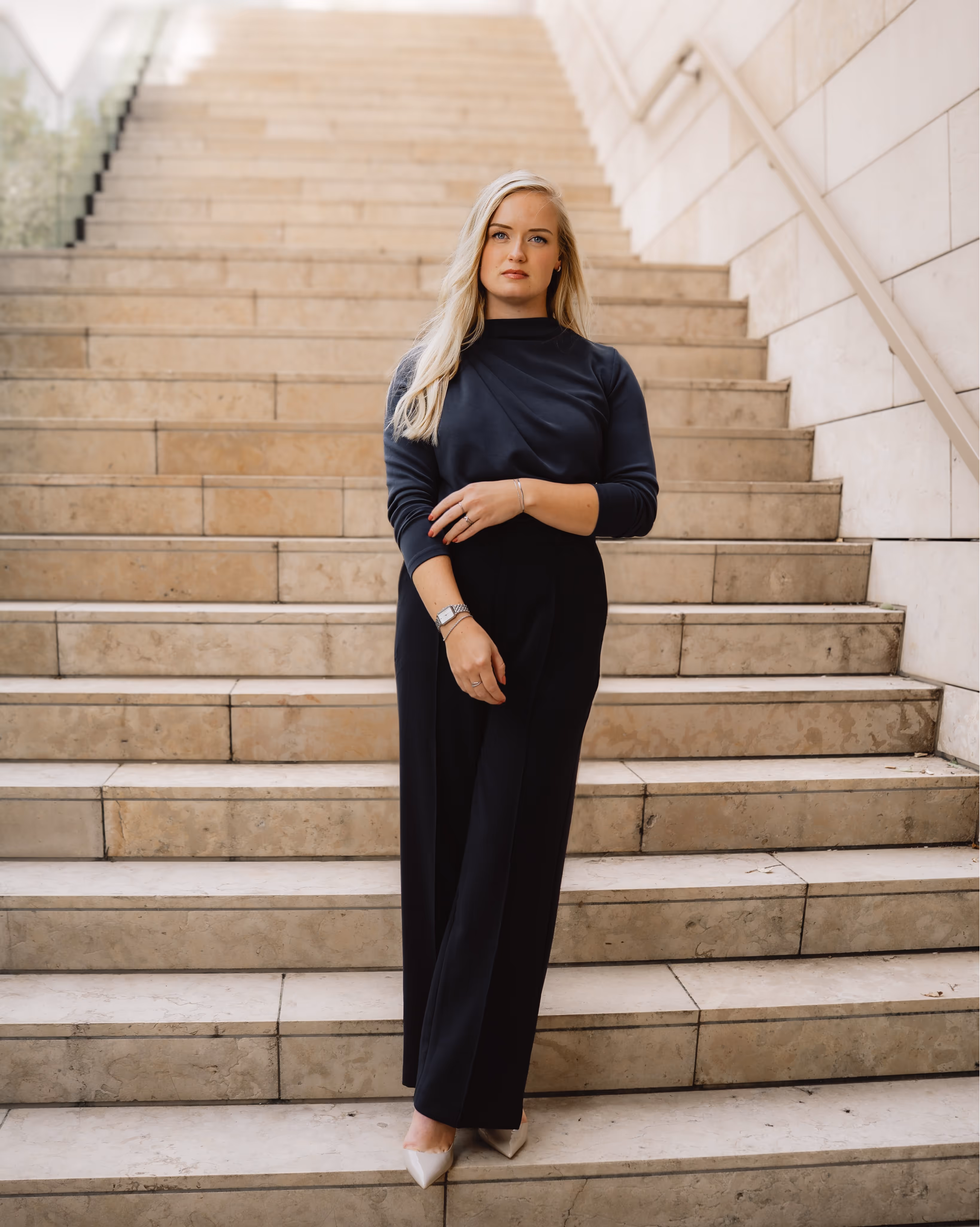 Vrouw met lang blond haar in een zwart ensemble en witte pumps staat op een brede stenen trap met een lichte muur aan de rechterkant.