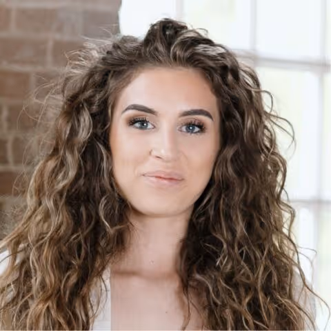 Vrouw met lang krullend haar en lichte huid, kijkend in de camera met een neutrale glimlach.