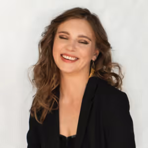 Vrouw met golvend kastanjebruin haar en zwarte blazer lacht met gesloten ogen tegen een lichte achtergrond.