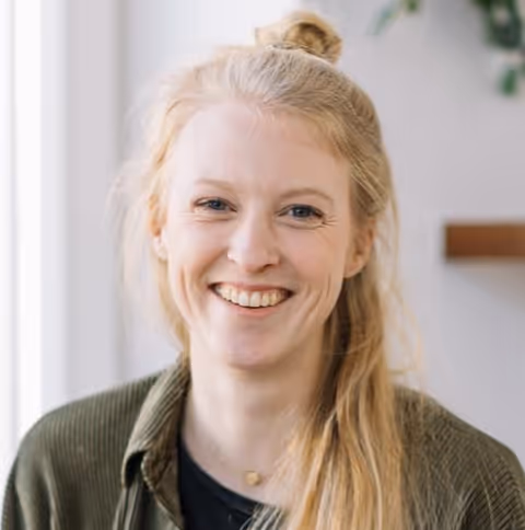 Lachende vrouw met lang, blond haar, groene blouse en een gouden hartvormige ketting.