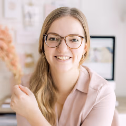 Vrouw met lang blond haar en bril glimlacht vriendelijk in een lichte kamer met een computer op de achtergrond.