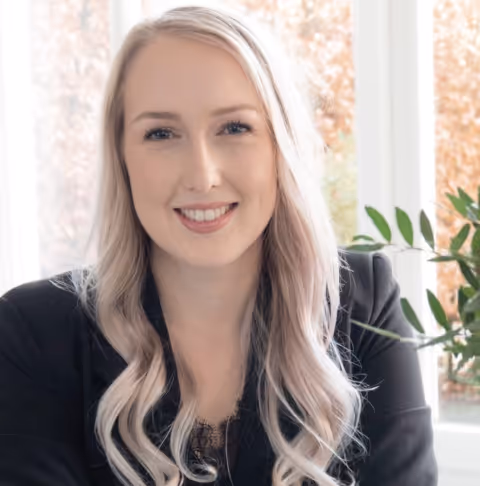 Blonde vrouw met golvend haar glimlacht en draagt een zwarte blazer, zittend voor een raam met herfstbladeren buiten.