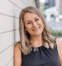 Vrouw met halflang blond haar in een zwart overhemd leunt tegen een witte muur en lacht naar de camera.