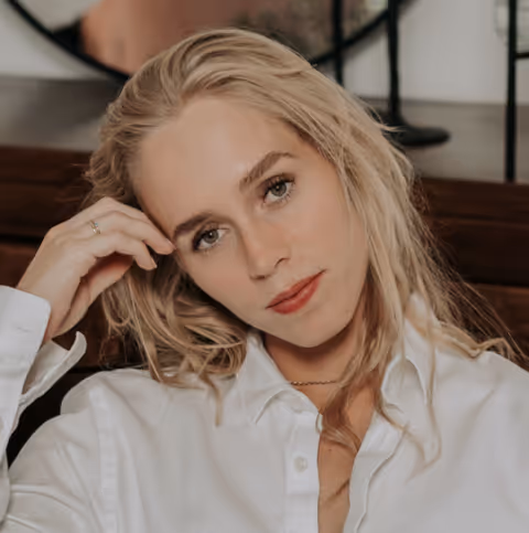 Blonde vrouw met lichte huid en losse haren, zittend en leunend met haar hand tegen haar hoofd, draagt een witte blouse.