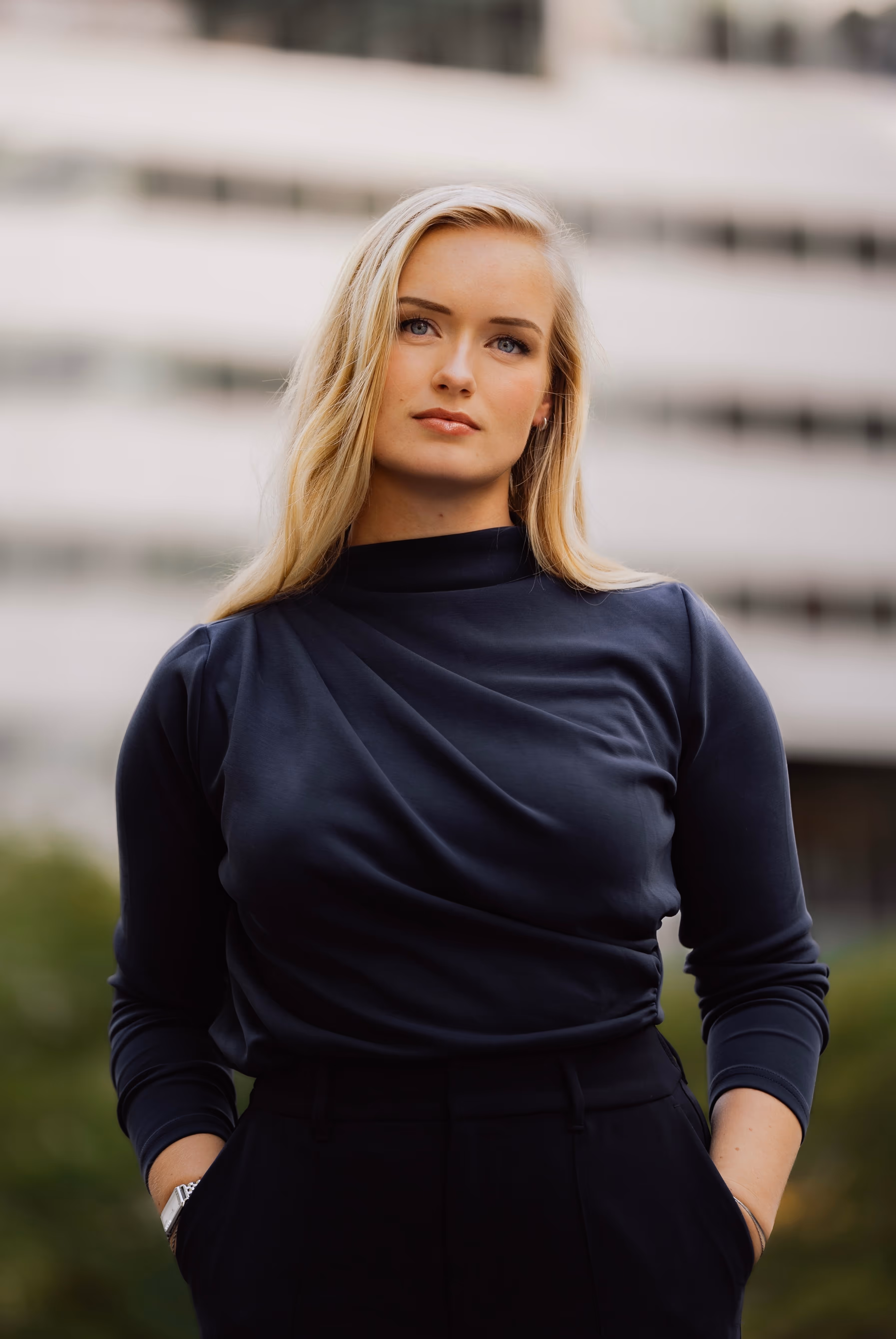 Tessa de Vries, een vrouw met lang blond haar en een donkere trui met lange mouwen, staat buiten met handen in de zakken.