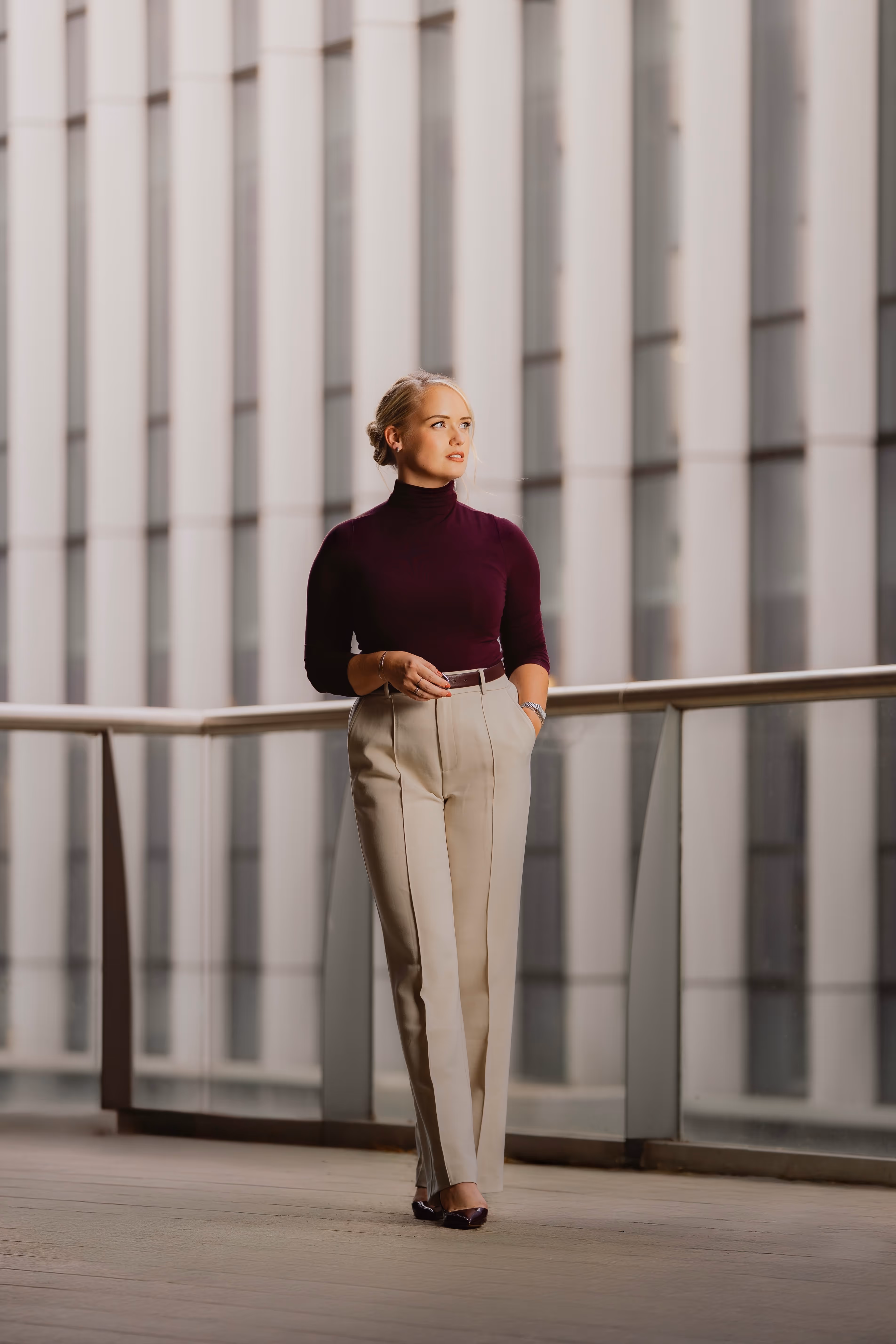 Tessa de Vries, een vrouw met beige broek en paarse coltrui staat op een balkon met glazen balustrade en kijkt naar rechts.