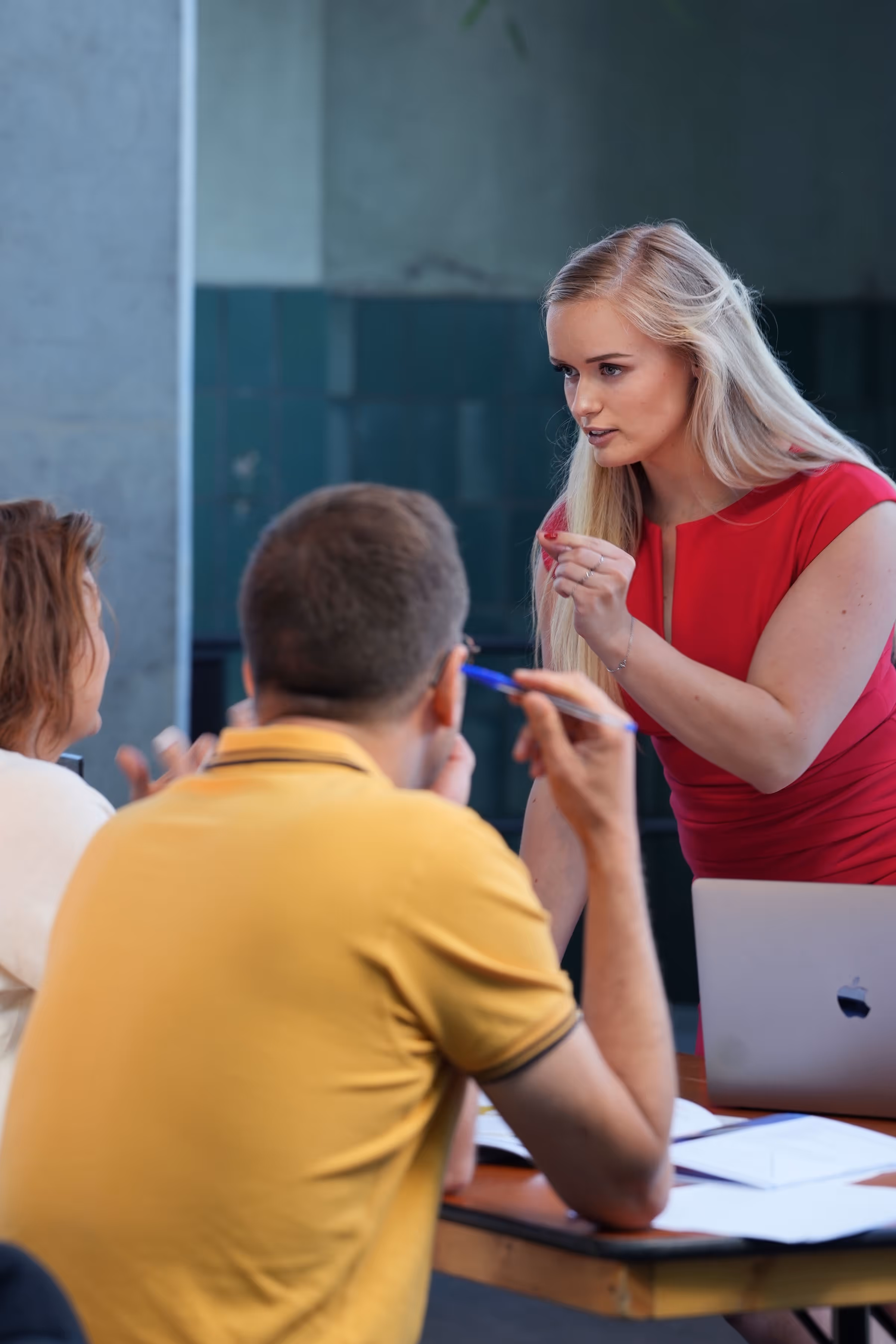 Tessa de Vries, een vrouw in een rood jurkje bespreekt iets serieus met een man en vrouw aan een tafel met een laptop en documenten.