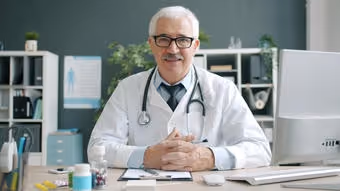 Smiling male doctor with glasses and stethoscope sitting at a desk in a medical office.