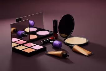 Assorted makeup products including powder compacts, lipstick, blush palette, and nail polish on a purple surface.
