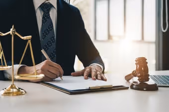 Person in suit writing on a document at desk with legal scales and judge's gavel nearby.