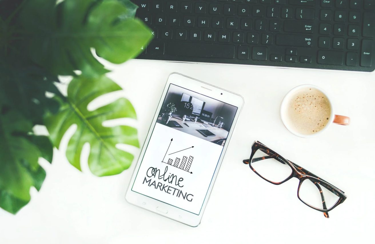 An iPad with 'online marketing' written on the screen, sunglasses, coffee, and a black keyboard on a white table