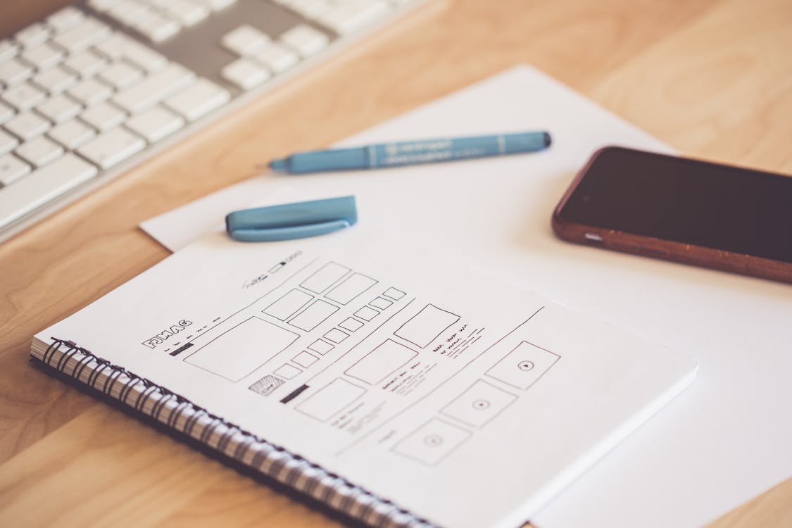 A UX Web Design Drawn on a Notebook with a Pen Lying on a Table Beside a Phone and Keyboard