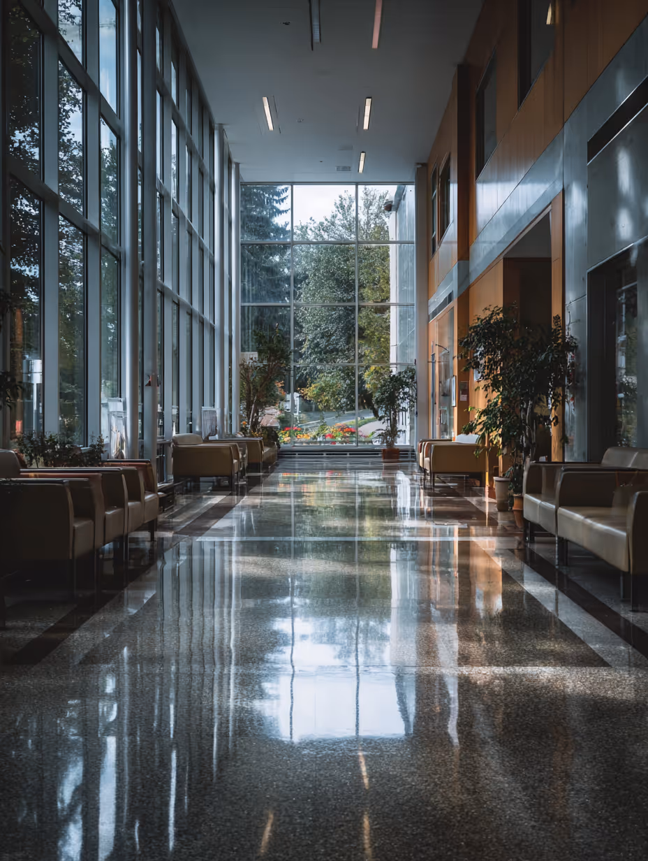 Bright corridor with large floor-to-ceiling windows on the left, reflecting on polished floor, brown armchairs lining both sides, and potted plants near a glass wall at the far end.