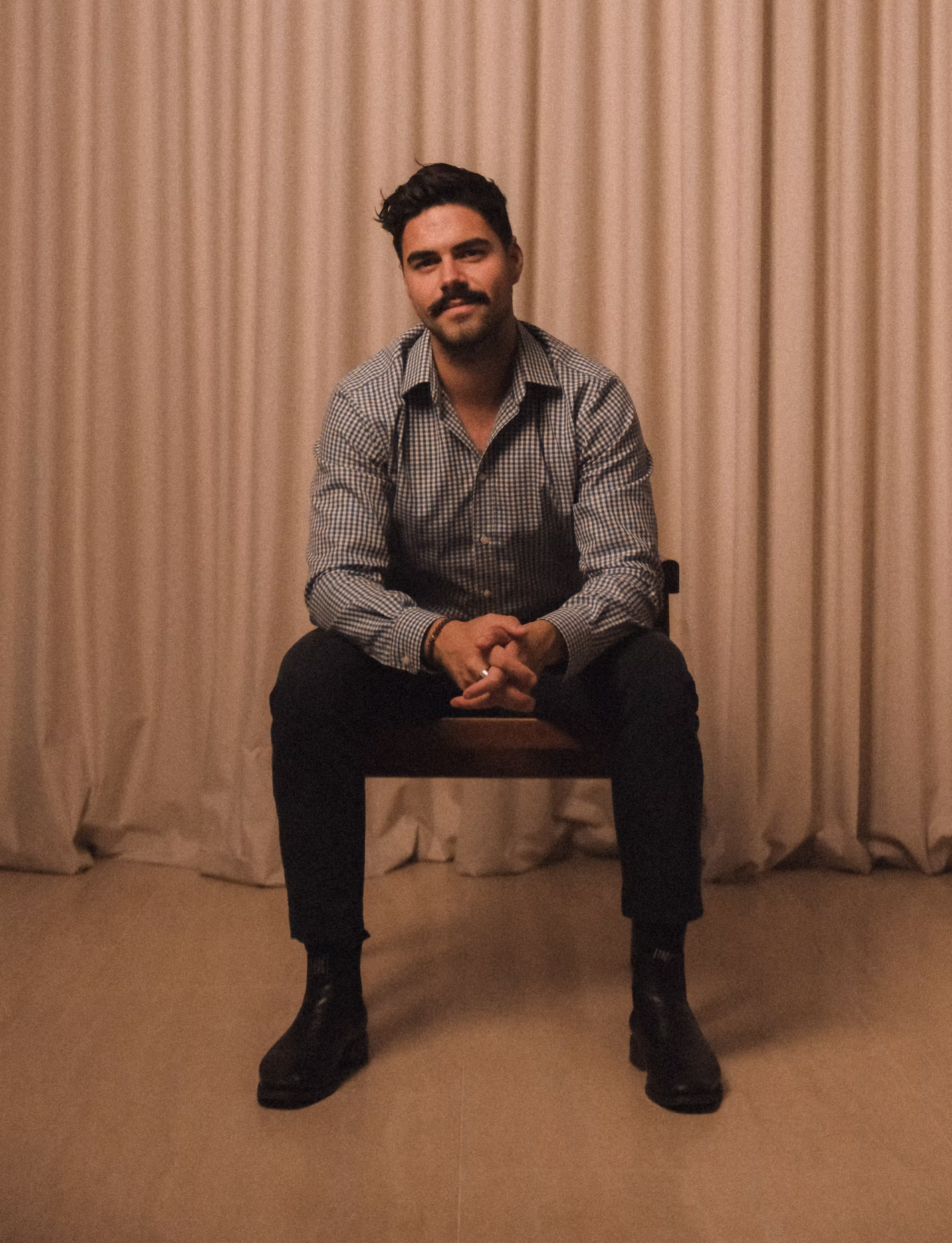 Hamish Caulfield sitting on a wooden chair, wearing a checkered shirt, black pants, and black boots against a beige curtain background.