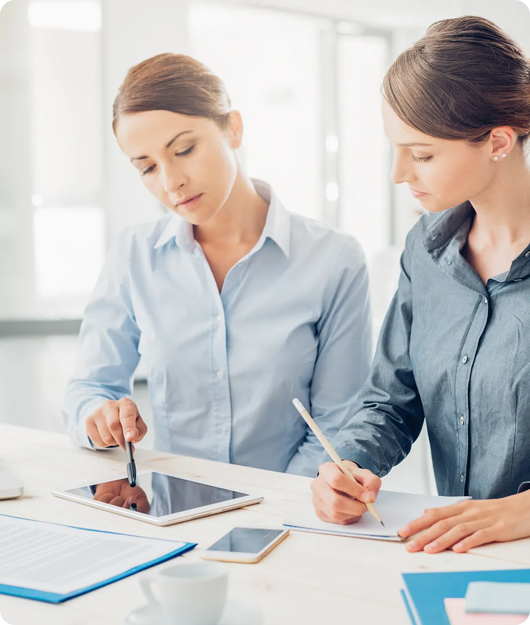 Two women in an office discussing work, one pointing at a tablet while the other writes notes on paper.