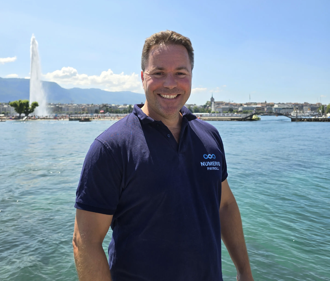 Ein lächelnder Mann in einem marineblauen Poloshirt mit dem Numeriq Payroll-Logo steht am Ufer, im Hintergrund sind die Skyline, ein Springbrunnen und ein strahlend blauer Himmel zu sehen.