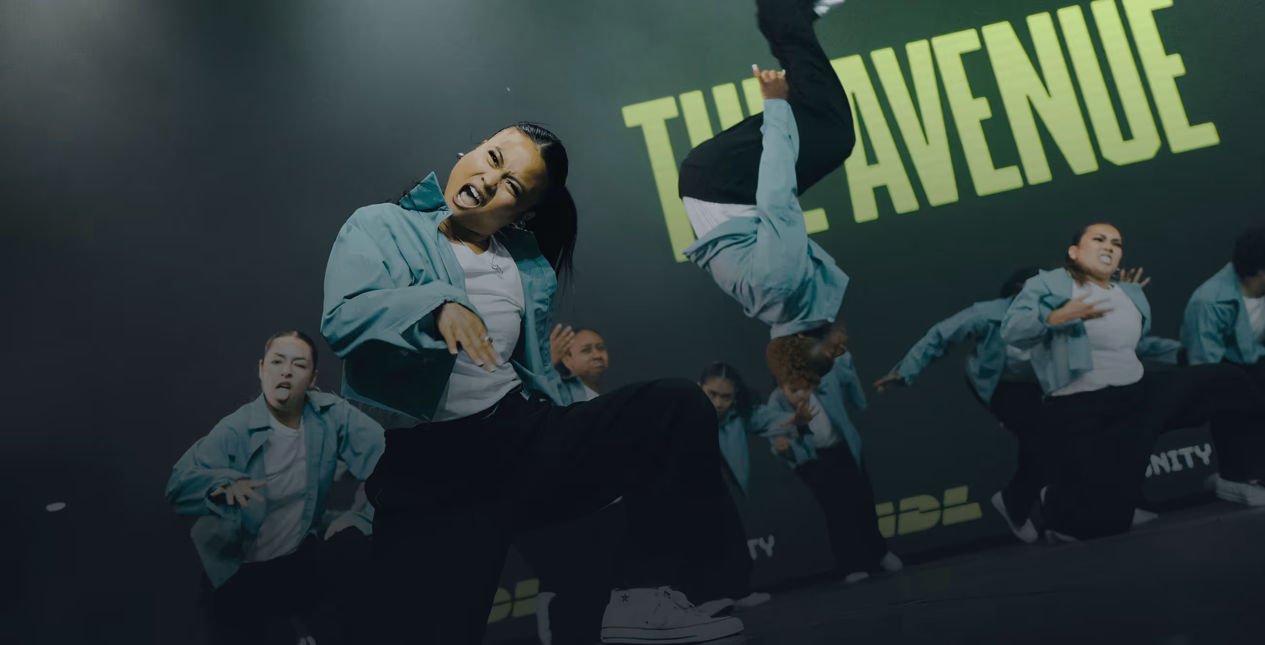 Group of dancers performing energetically on stage wearing teal jackets and black pants with a backdrop reading 'THE AVENUE'.