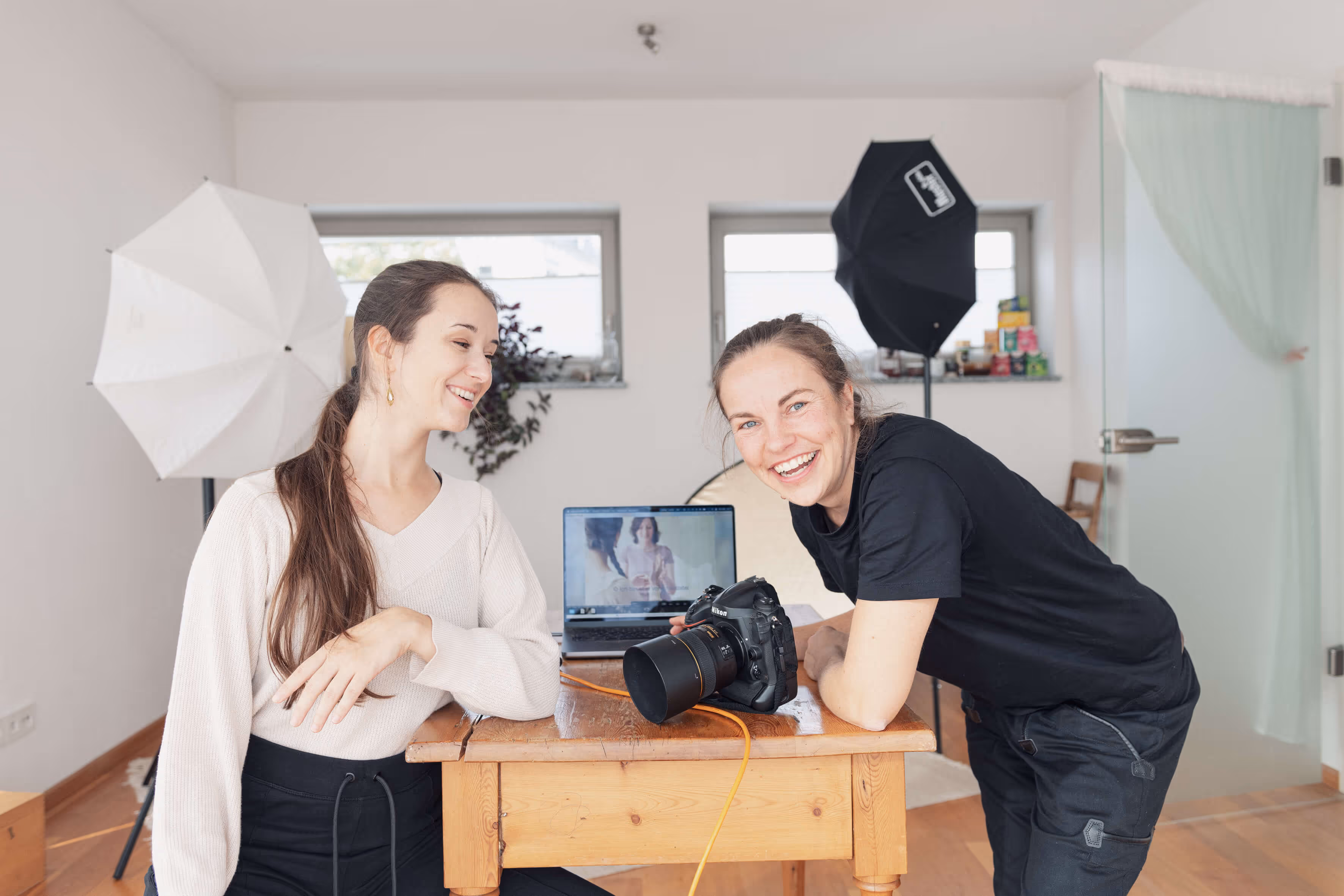 Anna Genov und eine Fotografin in einem Fotostudio lächeln und lehnen sich an einen Holztisch mit Kamera und Laptop.