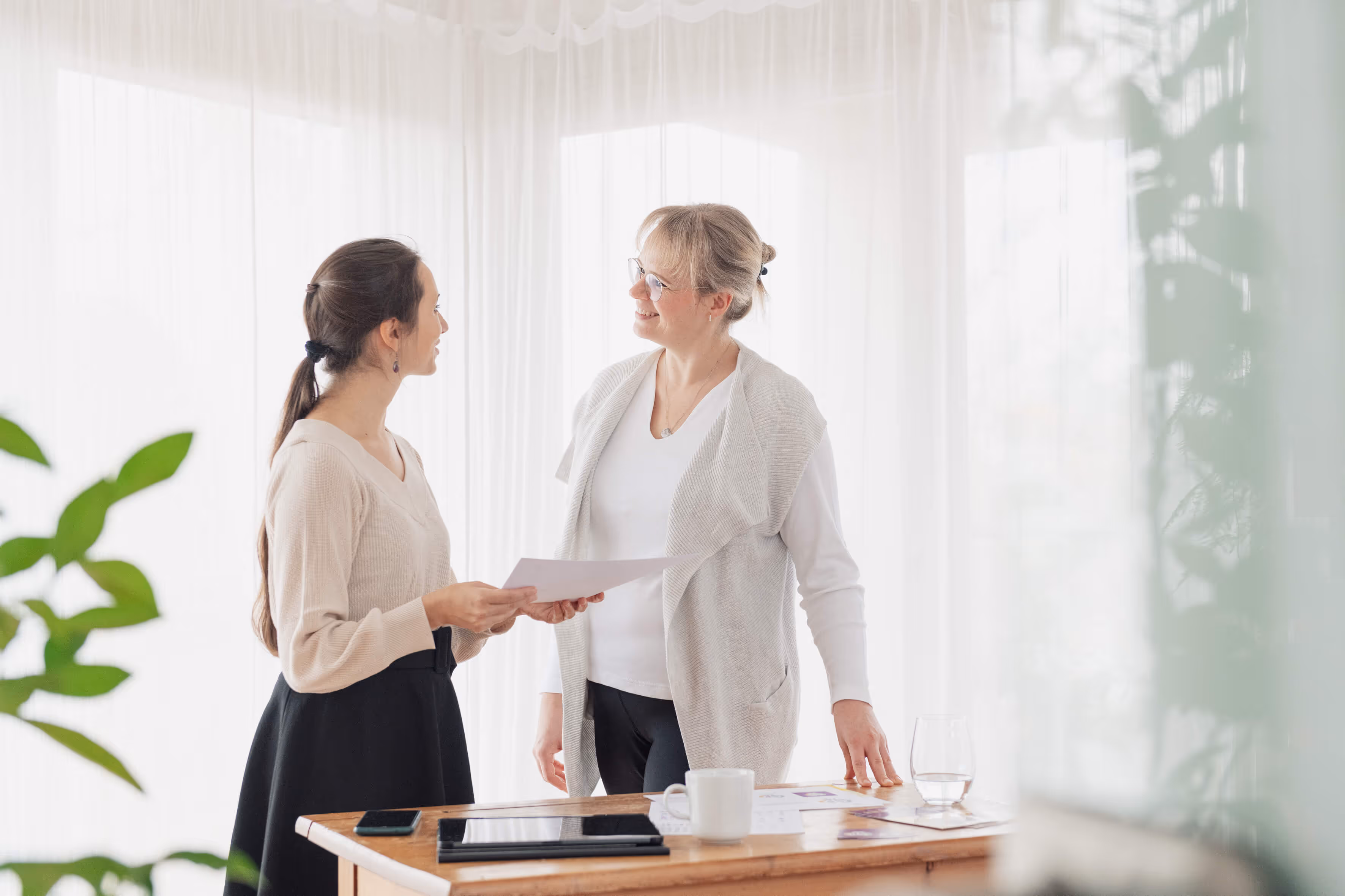 Anna Genov steht mit einer Kundin an einem Holzschreibtisch. Sie hält ein Blatt Papier in der Hand und unterhält sich mit der Kundin in einem hellen Raum mit weißen Vorhängen.