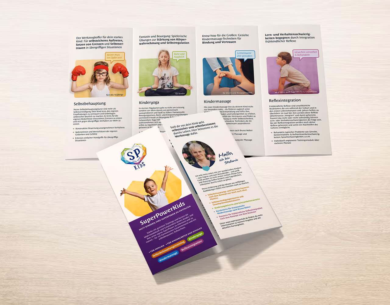 Drei aufgeklappte Broschüren liegen auf einem hellen Holztisch, zeigen Informationen und Fotos zu Selbstbehauptung, Kinderyoga, Kindermassage und Reflexintegration für Kinder. Zwei weitere Broschüren sind geschlossen, eine mit dem Titel SuperPowerKids und einem lachenden Kind auf der Vorderseite.