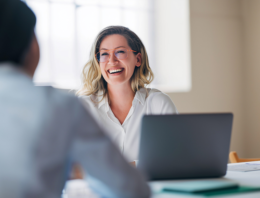 Smiling woman with glasses sitting at a desk with a laptop, talking to another person.