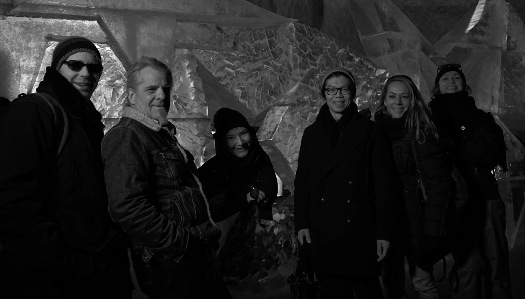 Group of six adults in winter clothing standing indoors in front of a textured wall, smiling at the camera.