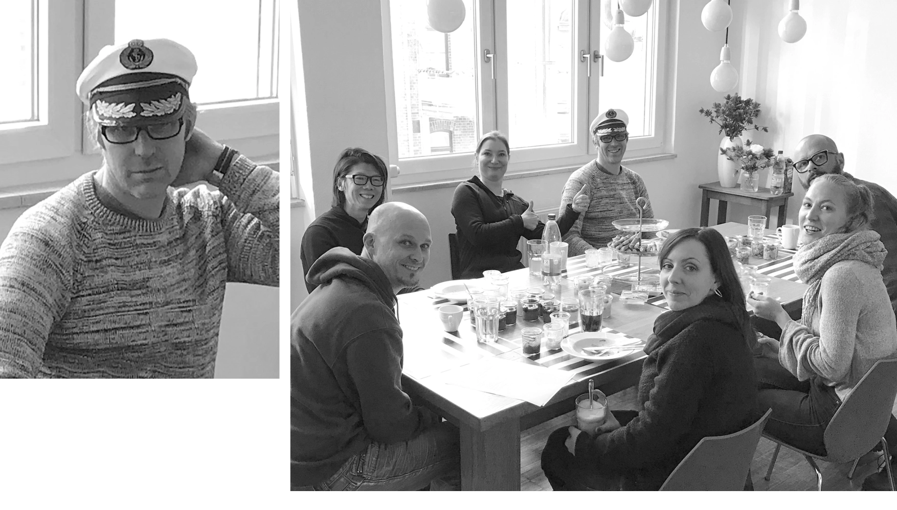 Group of eight people sitting around a table with drinks and snacks, two wearing captain hats, giving thumbs up and smiling indoors.