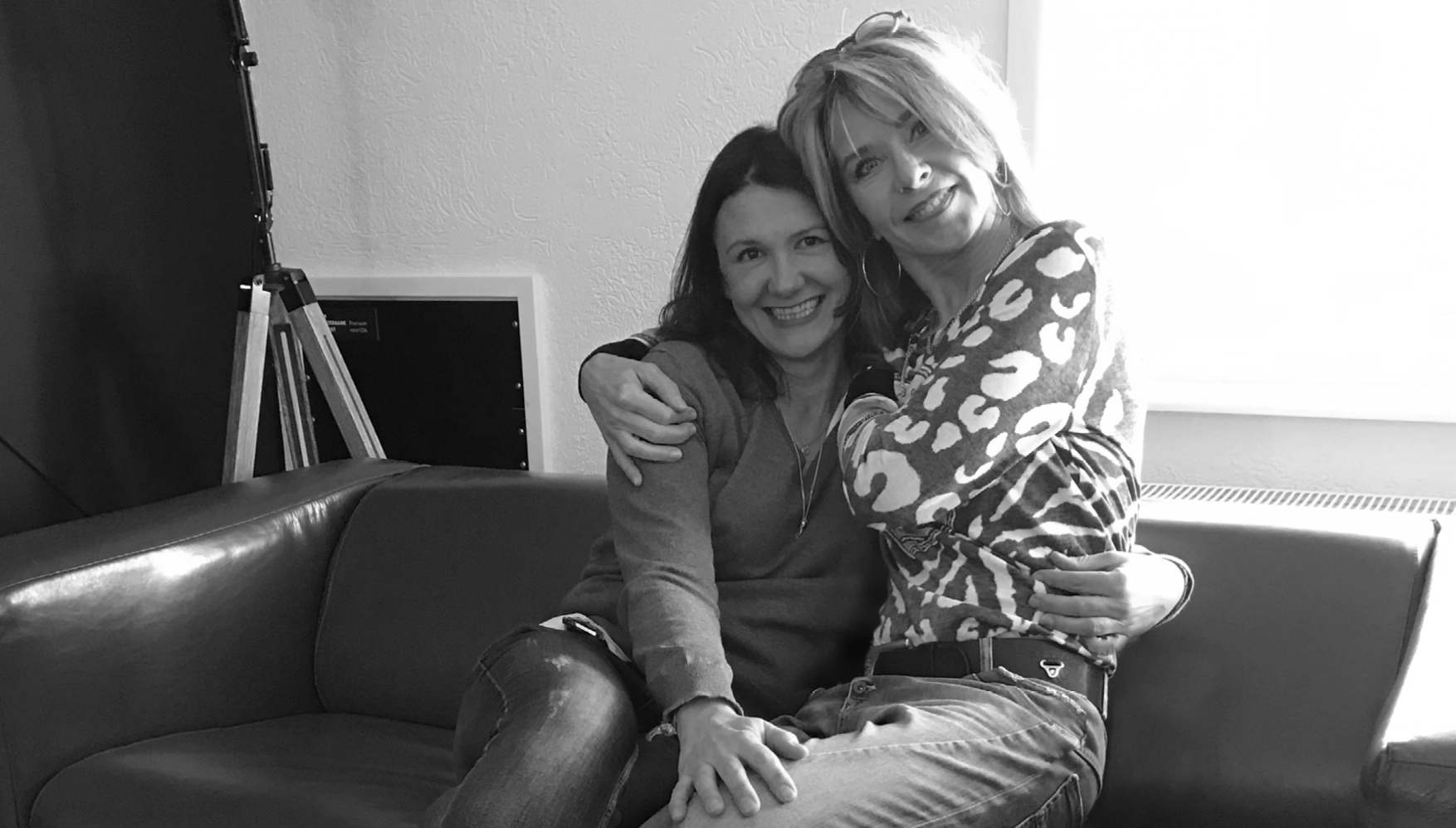 Two women sitting closely on a couch, smiling and hugging each other warmly.