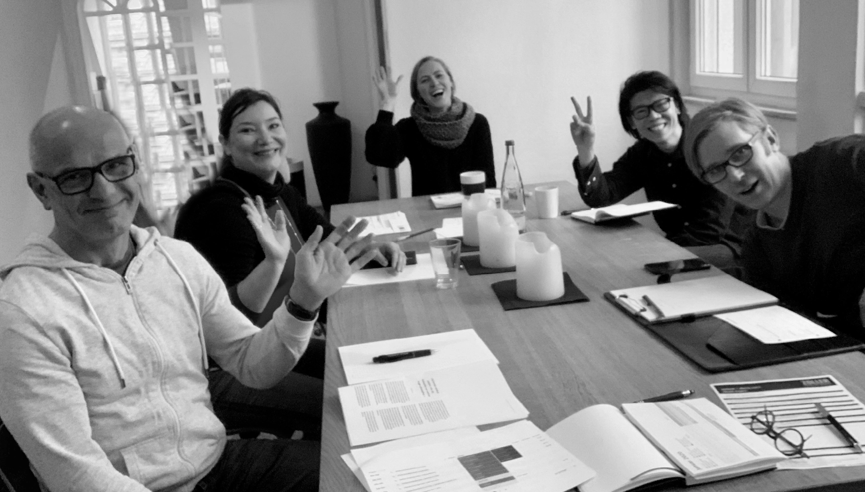 Five people sitting around a table with documents, smiling, waving and making peace signs in a bright meeting room.